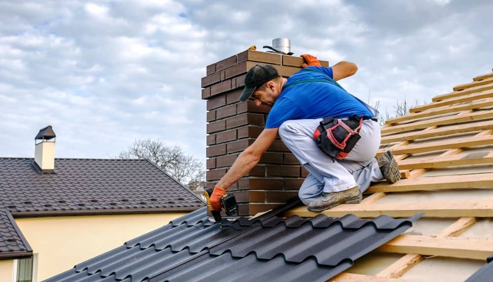 Construction worker on a sloped roof installing dark metal roofing panels beside a brick chimney, wearing a tool belt and gloves with an orange glove on one hand.